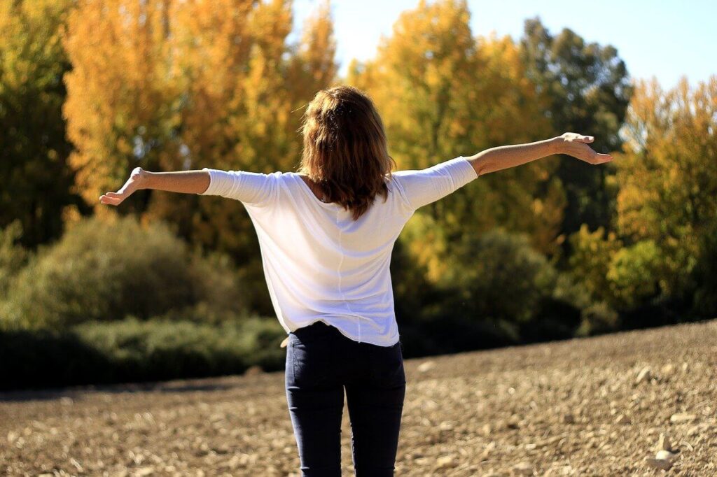 Woman in a white blouse stands with her back turned and arms outstretched in front of autumn trees