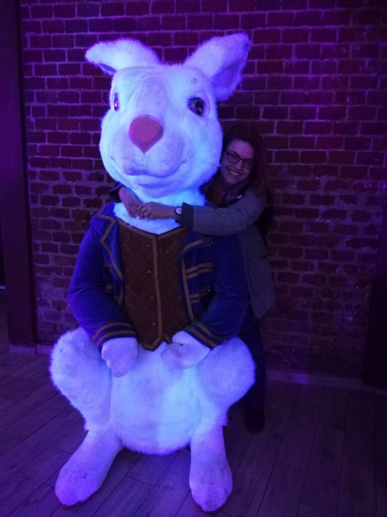 Kobieta w okularach obejmuje dużą postać białego królika w kamizelce przy ceglanej ścianie