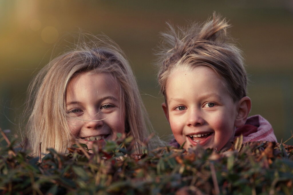Two smiling children peeking out from behind a bush