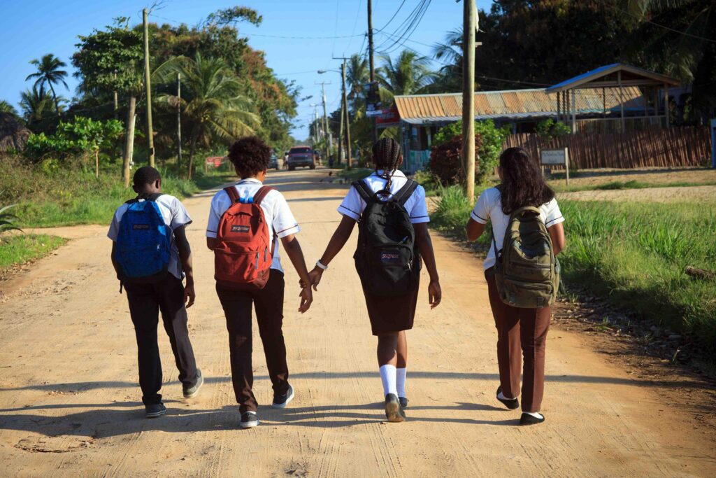 Four schoolchildren in uniforms with backpacks walking along a sandy path among palm trees