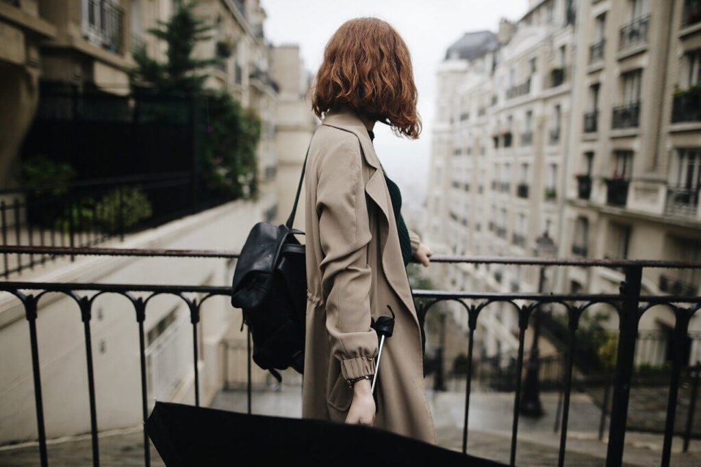 Red haired woman in a coat with a backpack stands by a railing between tenement buildings