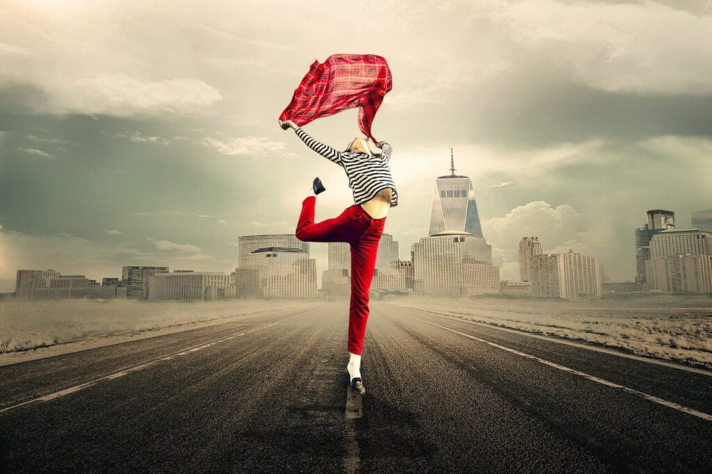 Woman in red trousers dances with a red scarf on an empty road in front of high rise buildings